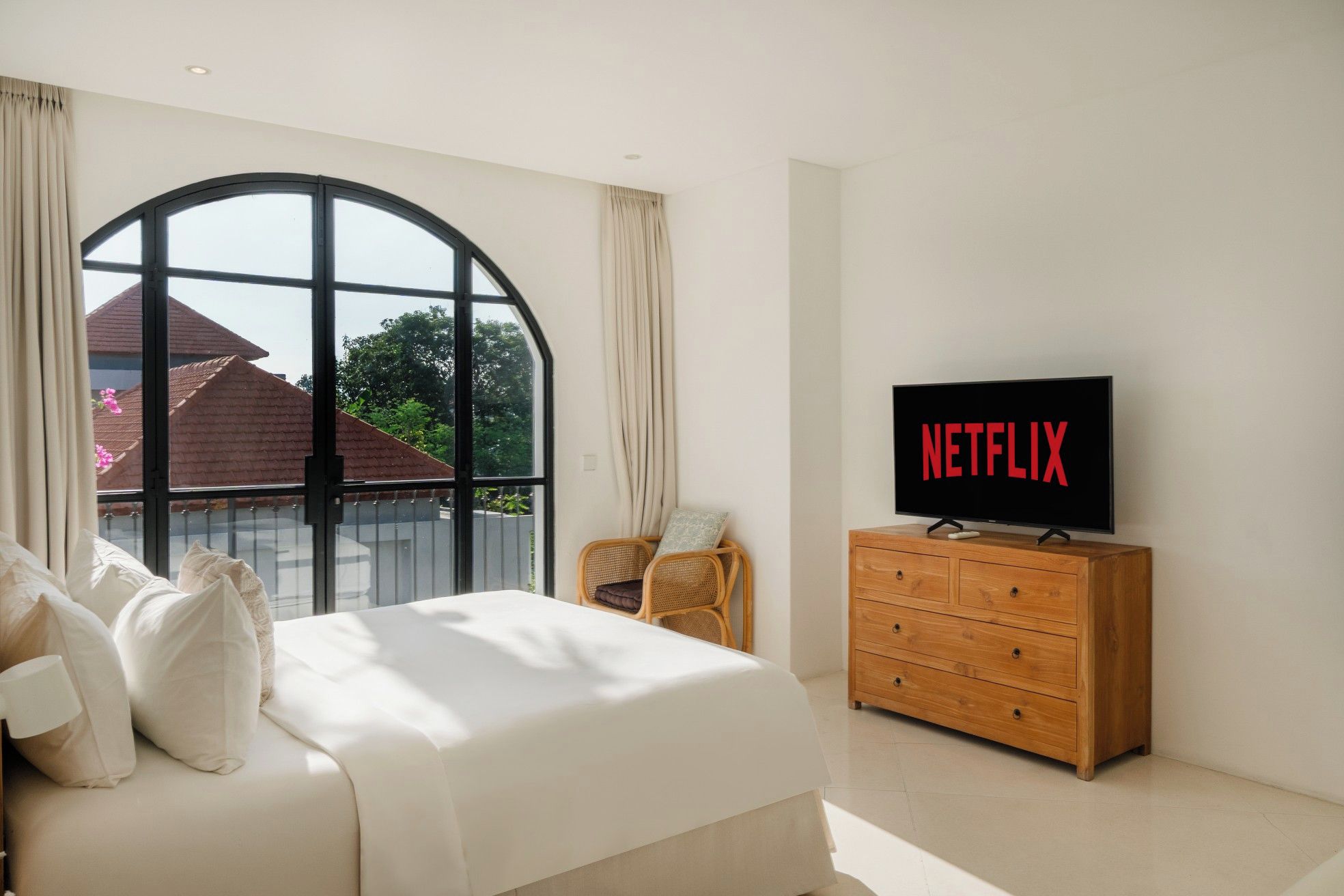 A bedroom with a large window, a bed with pillows, a wooden dresser, and a television displaying the Netflix logo.