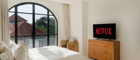 A bedroom with a large window, a bed with pillows, a wooden dresser, and a television displaying the Netflix logo.