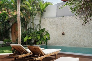 Un espace aménagé au bord de la piscine, avec des chaises longues, un mur en pierre et des plantes tropicales.