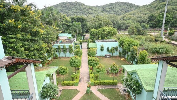 A courtyard with a central pathway, surrounded by green lawns and trees, leading to a building with a green roof. - Ranthambhore Vatika Resort (Sawai Madhopur)
