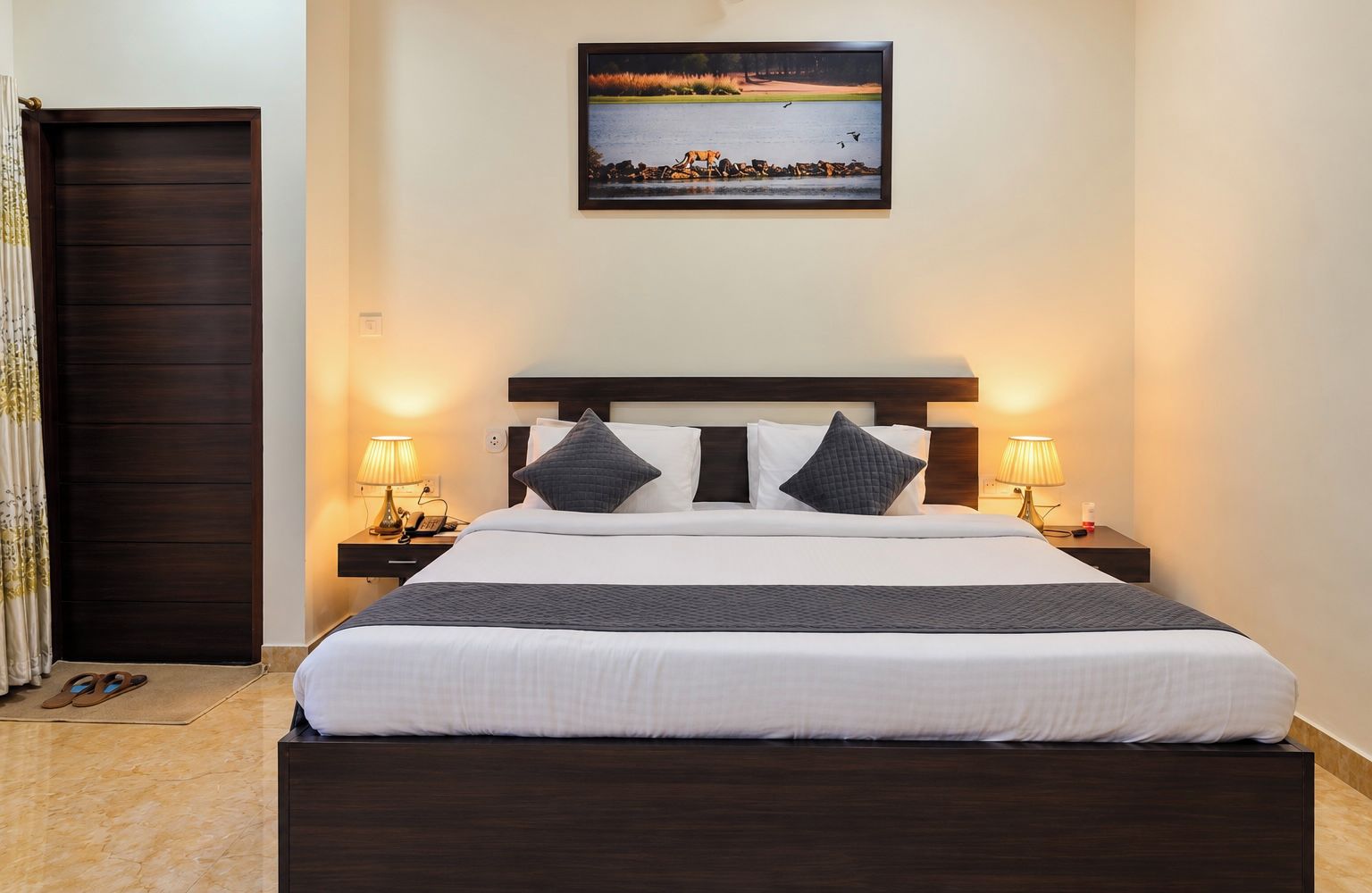 A hotel room with a large bed, two bedside tables, a dark wooden headboard, a framed landscape painting above the bed, and a dark wooden door.