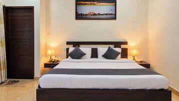 A hotel room with a large bed, two bedside tables, a dark wooden headboard, a framed landscape painting above the bed, and a dark wooden door.