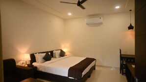 A hotel room with a bed, bedside lamps, a ceiling fan, an air conditioning unit, and a small table with a chair. - Ranthambhore Vatika Resort (Sawai Madhopur)