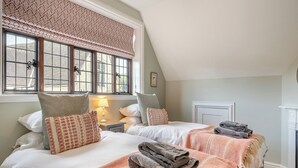 A bedroom with two beds, a window with a patterned valance, a bedside table with a lamp, and a door. - 2 bedroom accommodation in Sturminster Newton (Sturminster Newton)