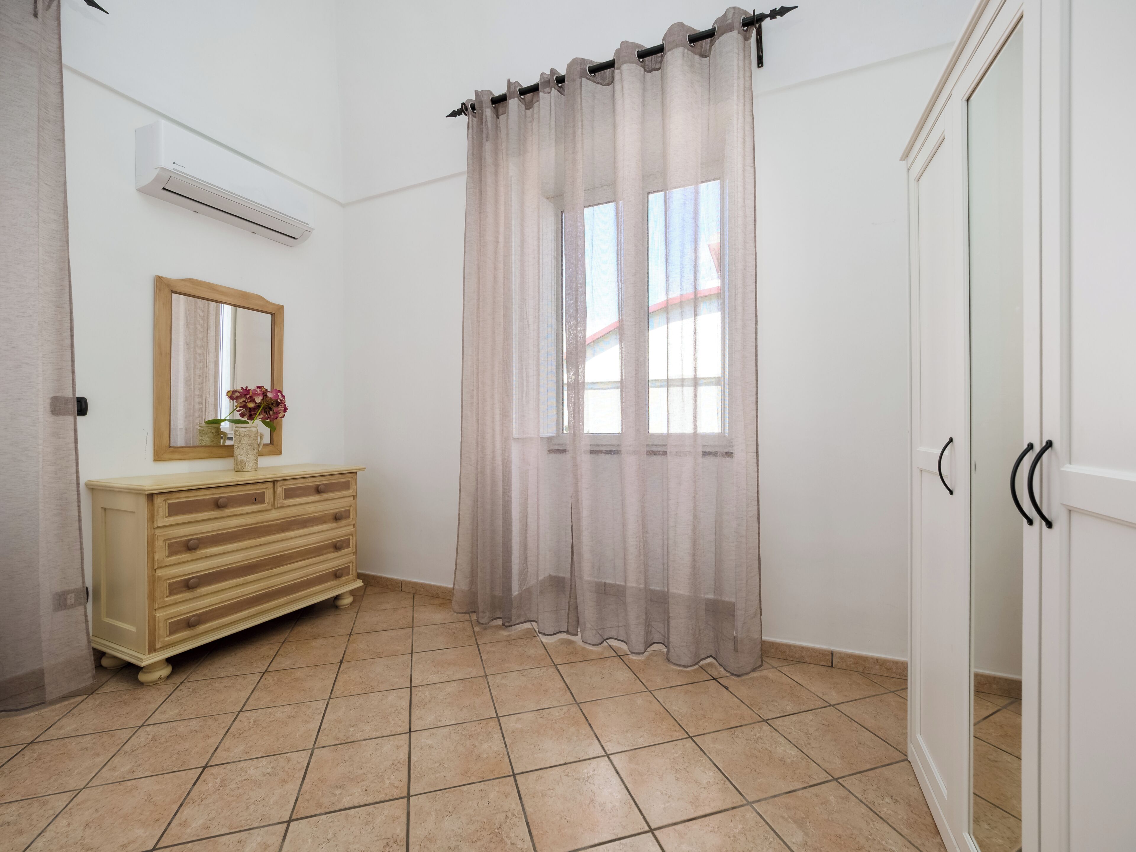 A bathroom with a beige tiled floor, a wooden vanity with drawers, a mirror, a window with sheer curtains, and a white door.