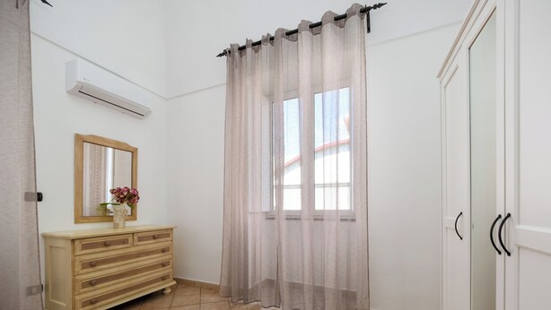 Ein Badezimmer mit beigefarbenen Fliesen, einem Holzkosmetikschrank mit Schubladen, einem Spiegel, einem Fenster mit durchsichtigen Vorhängen und einer weißen Tür.