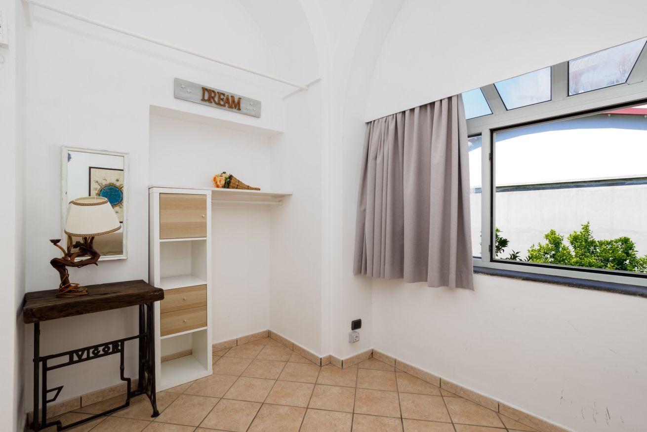 Um quarto com parede branca, mesa consola de madeira, estante com objetos decorativos, janela com cortinas e um letreiro que diz 'DRREAM'.