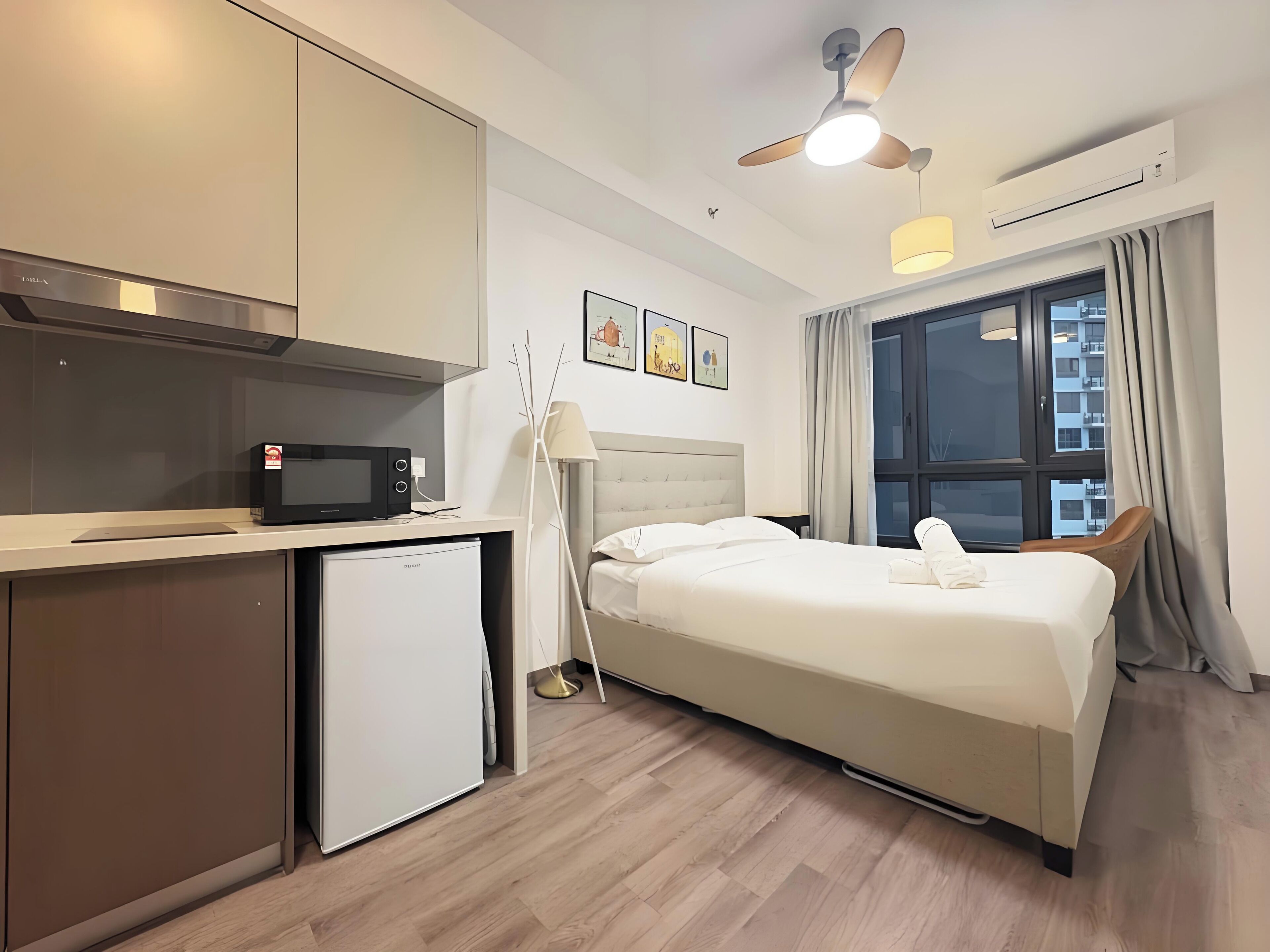 Habitación de hotel con cama, escritorio con microondas, refrigerador miniatura, ventilador de techo y ventana con cortinas.