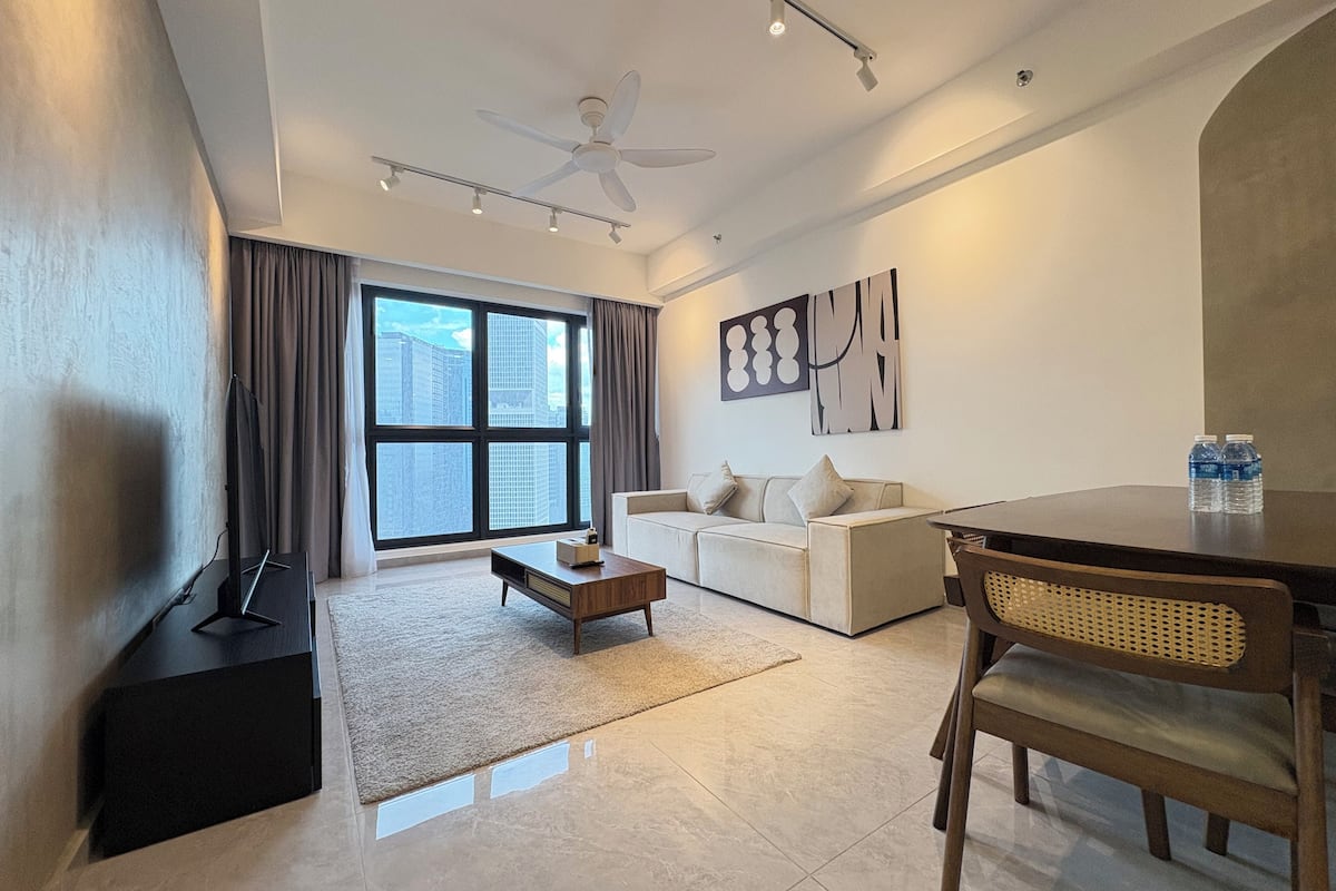 A modern living room with a white sofa, a coffee table, a dining table with chairs, a ceiling fan, and a large window with curtains.