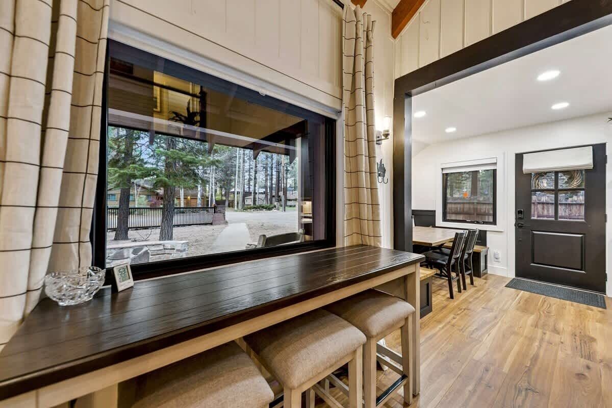 Una habitación moderna con una mesa de comedor de madera, taburetes de bar, un ventanal con vistas a árboles y una puerta oscura.