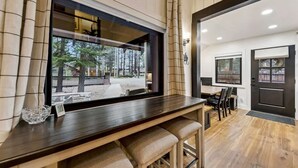 Una camera moderna con un tavolo da pranzo in legno, sgabelli da bar, un'ampia finestra con vista sugli alberi e una porta scura.