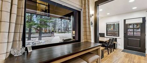 Una habitación moderna con una mesa de comedor de madera, taburetes de bar, un ventanal con vistas a árboles y una puerta oscura.