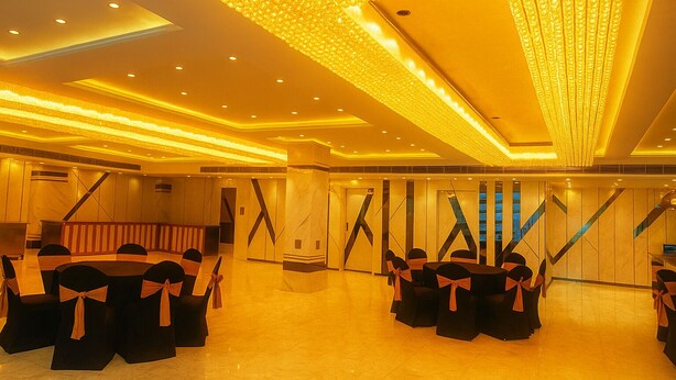 Une salle de banquet spacieuse, dotée d’un plafond haut, d’un éclairage encastré et d’un lustre central. Les tables sont disposées et les chaises noires sont ornées de rubans orange.