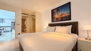Ein Hotelzimmer mit einem großen Bett, einem Nachttisch mit Lampe, einem Sonnenuntergangsbild und Blick auf das Badezimmer und die Küche durch eine offene Tür.