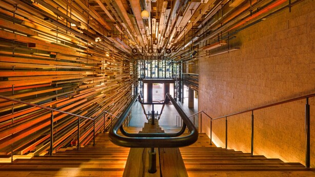 Un interior moderno con una escalera central flanqueada por vigas de madera y barandales de metal.