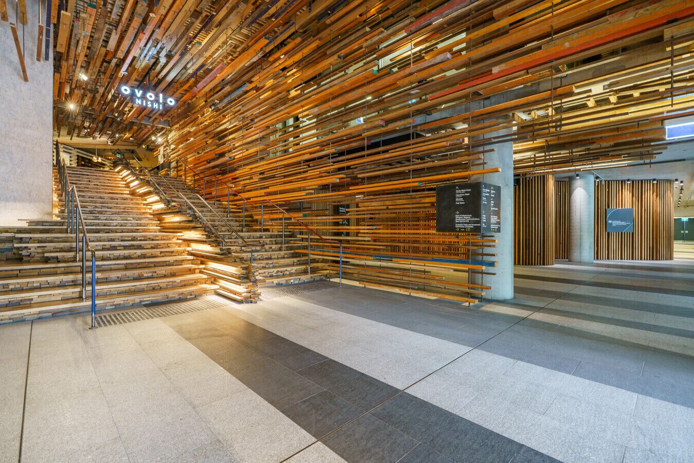 A modern interior with a staircase made of wooden planks, a sign reading "ovolo NISHI", and a tiled floor.