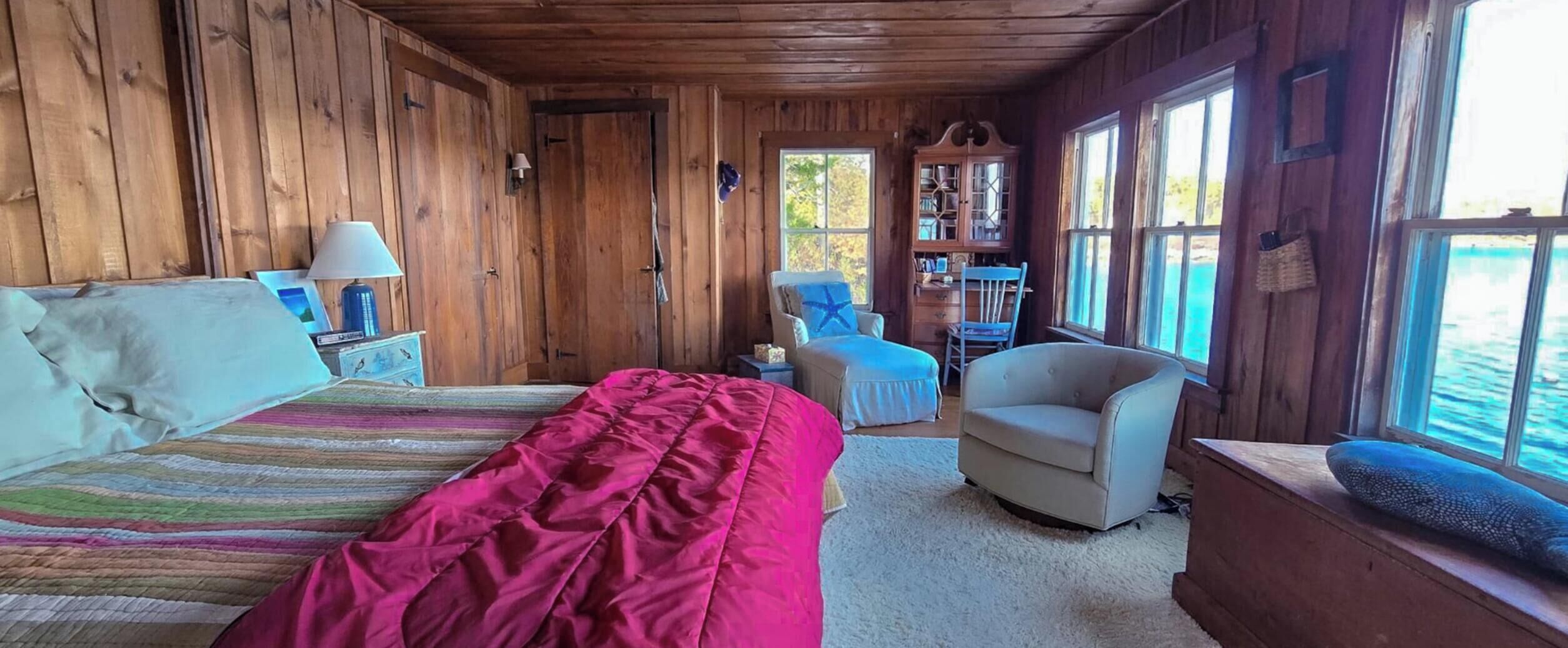 A cabin with a bed, a chair, a desk, and a window overlooking water.
