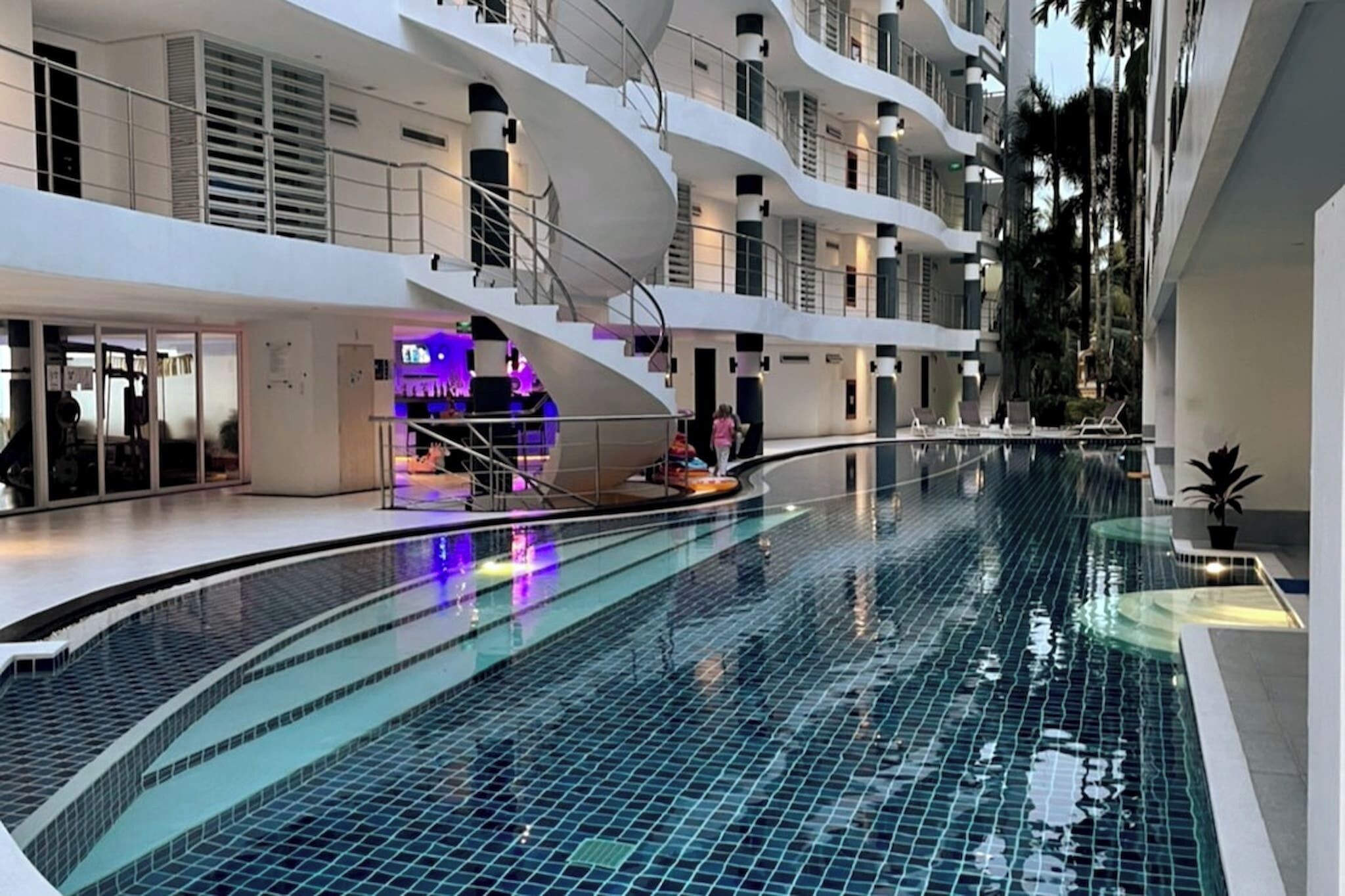 Ein moderner, innenliegender Schwimmbereich mit gebogener Form, umgeben von mehrstöckigen Balkonen und einer zentralen Treppe.