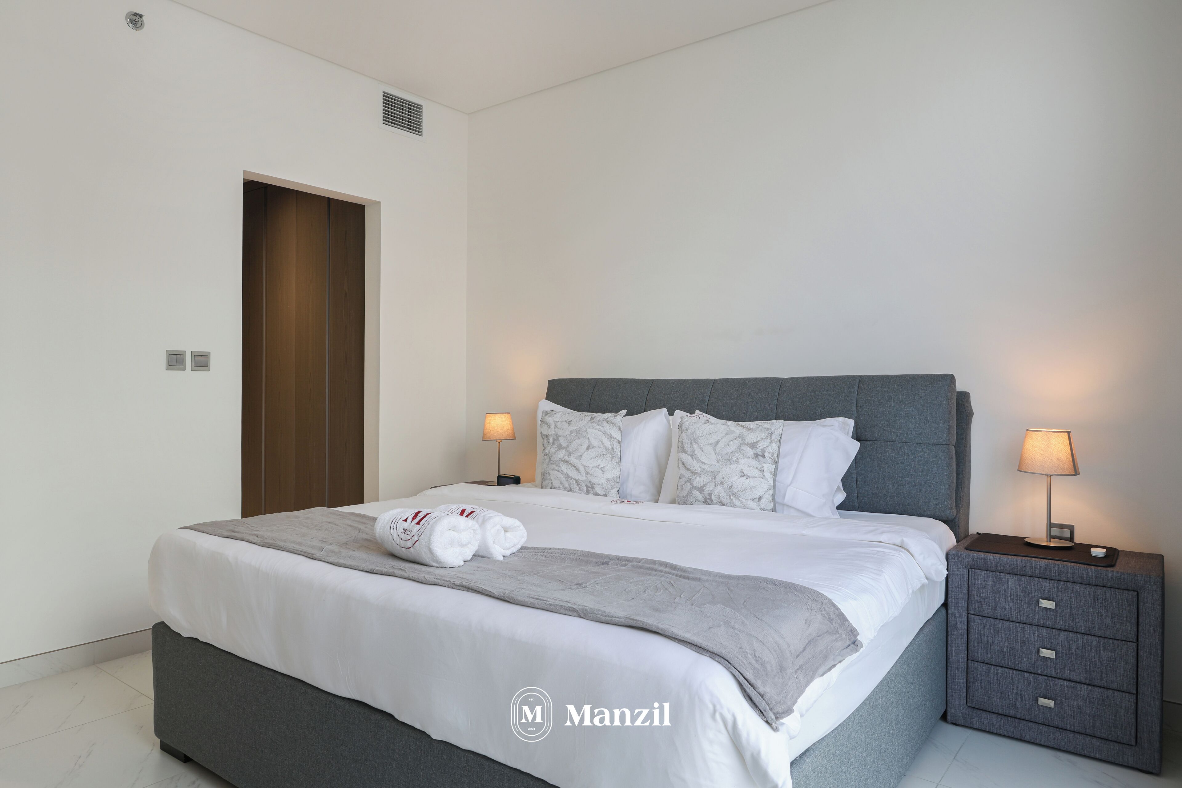 A modern bedroom with a large bed, grey bedding, and a nightstand with a lamp.