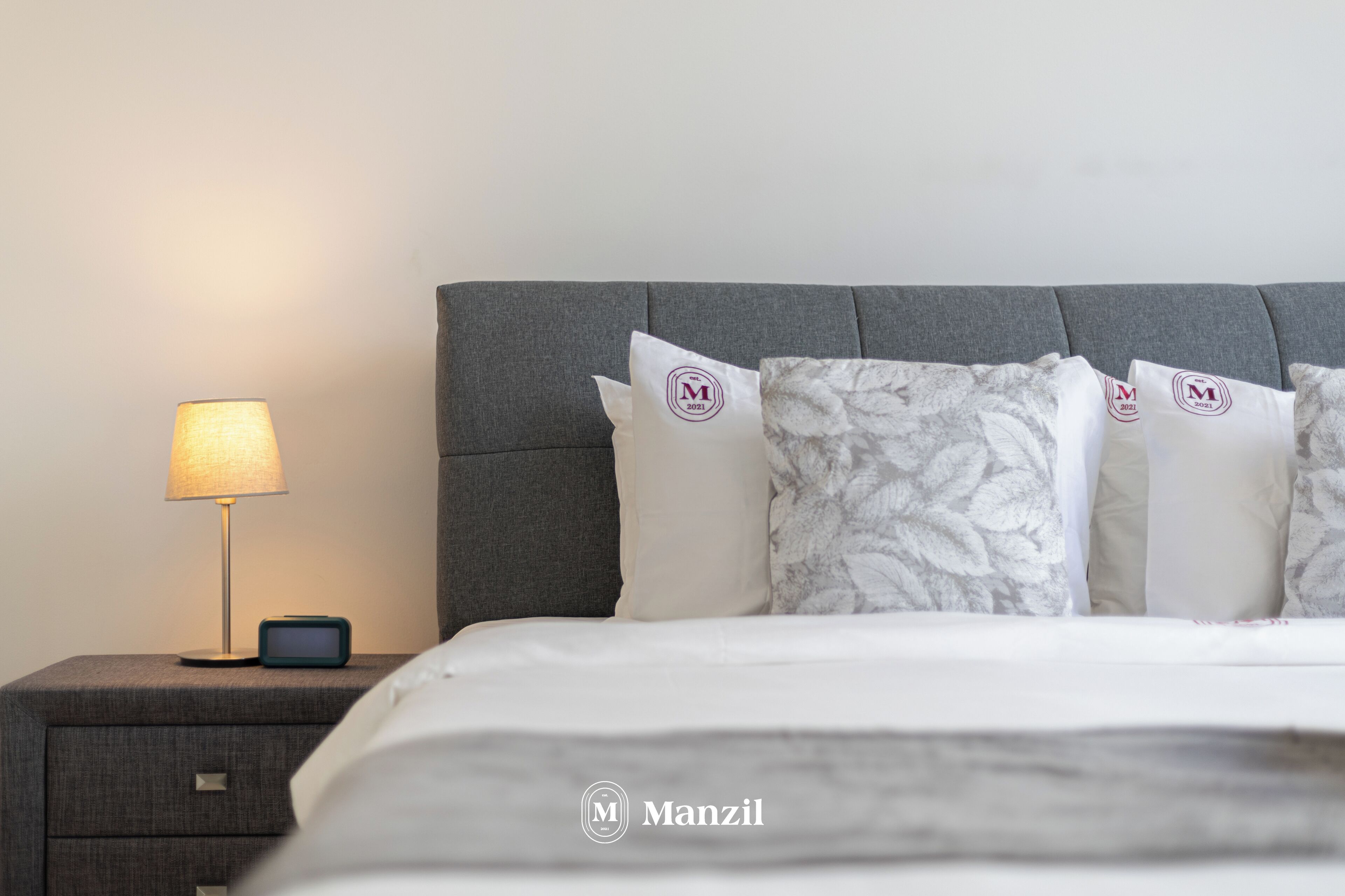 Una cama bien hecha con tablero cabecera gris, una lámpara de noche y una mesita de noche con un reloj.