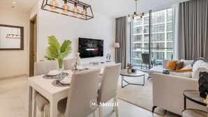 A modern living room with a sofa, dining table, and chairs, a flat-screen TV, and large windows with curtains.