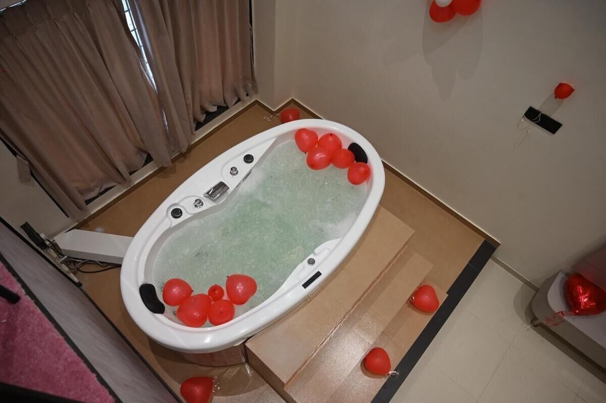 A bathtub filled with red balloons, surrounded by a wooden floor and beige curtains.