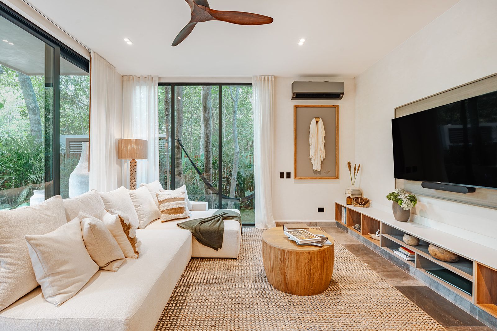 Una sala moderna con un televisor de pantalla plana grande, un sofá, una mesa de centro y vistas al exterior a través de amplios ventanales.