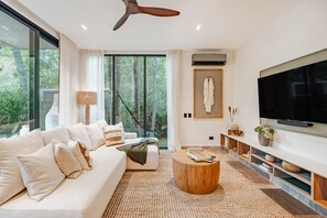 A modern living room with a large flat-screen TV, a sofa, a coffee table, and a view of the outdoors through large windows. - Kachava Lux (Tulum)