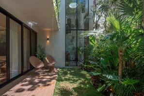 Un espacio exterior moderno con pared de vidrio, dos sillones de mimbre y exuberante vegetación tropical.