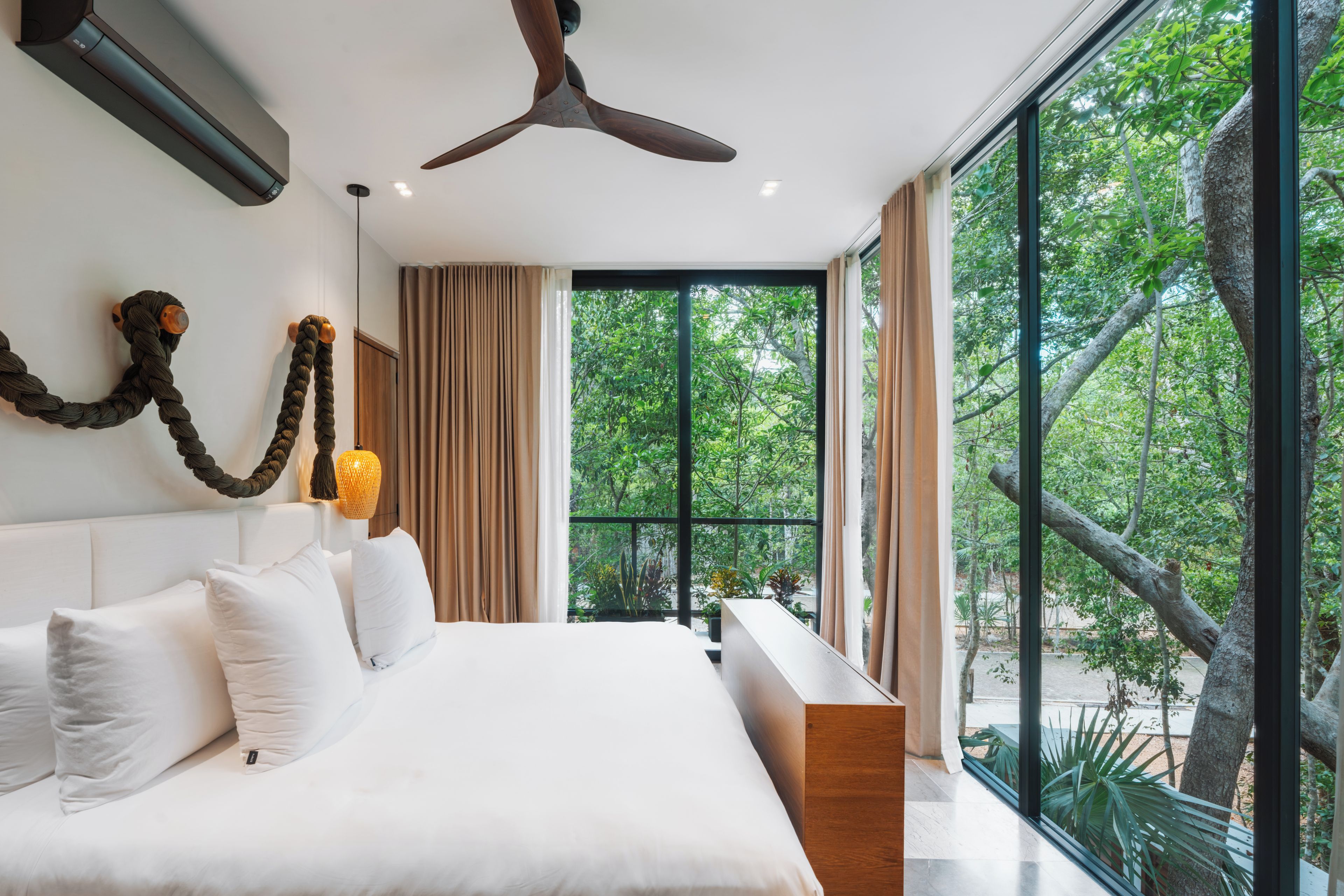 A modern bedroom with a large bed, a ceiling fan, and a view of greenery through large windows.