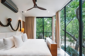 A modern bedroom with a large bed, a ceiling fan, and a view of greenery through large windows. - Kachava Lux (Tulum)