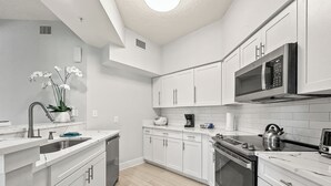 Una cocina moderna con armarios blancos, un lavamanos de acero inoxidable, un microondas y un hervidor en la estufa.