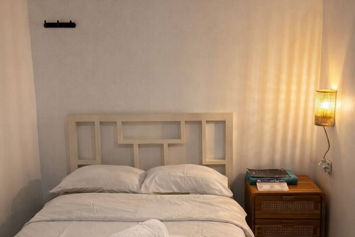 A neatly made bed with a rolled towel, a wooden nightstand with a lamp, and a wall-mounted shelf.