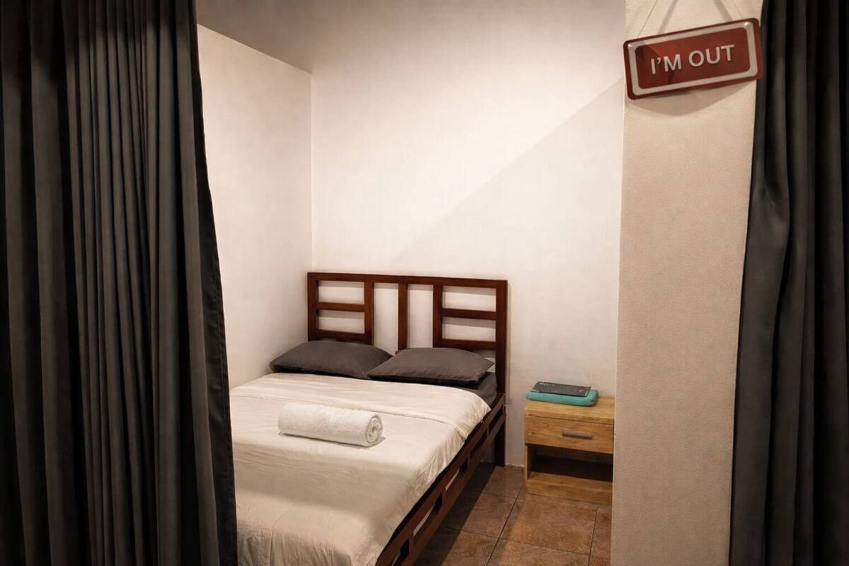 A small, neatly made bed with a wooden headboard and two pillows, a small bedside table with a book, and a sign on the wall that says "I'M OUT".