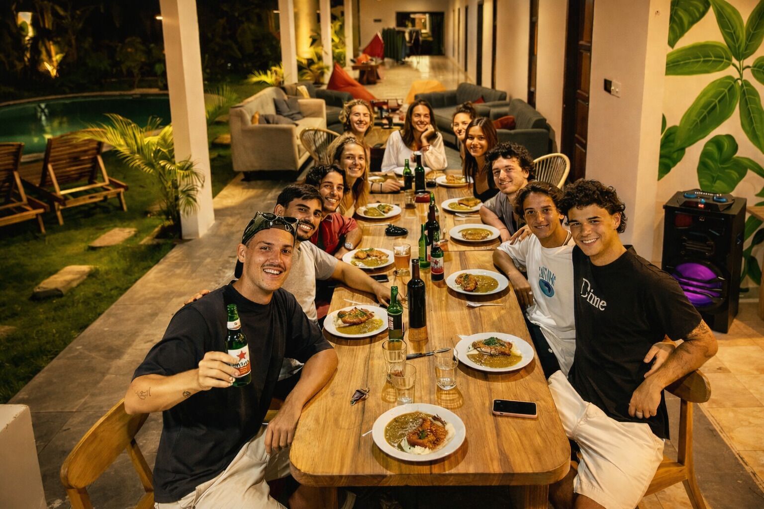 A group of people seated around a wooden dining table in an outdoor setting with a pool and greenery in the background.