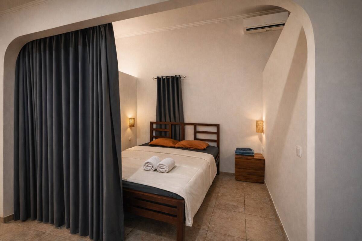A bedroom with a bed, dark curtains, two bedside lamps, and an air conditioning unit.