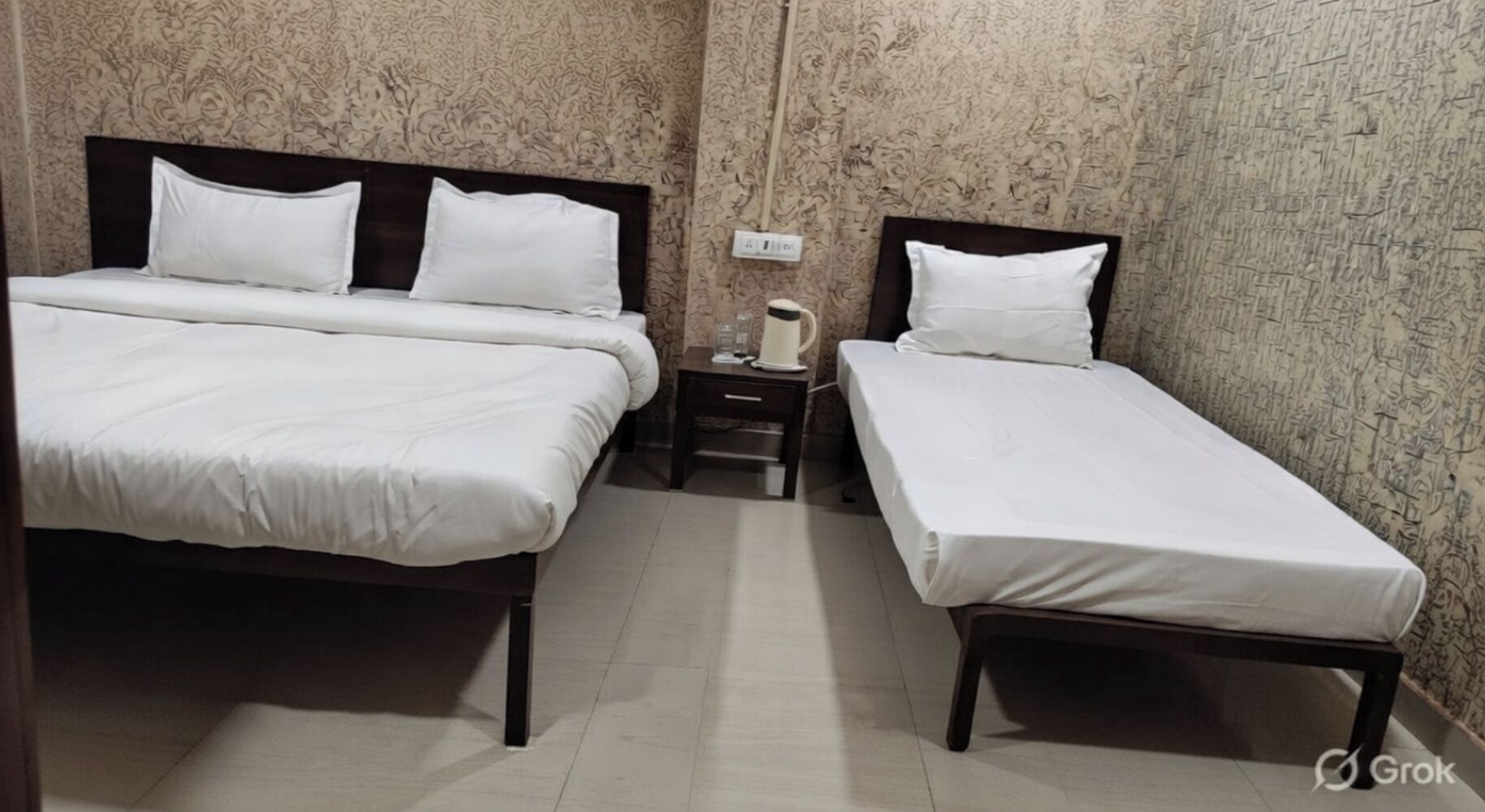 Een kamer met twee bedden, een nachtkastje met waterkoker en een muur met een bloemendesign.