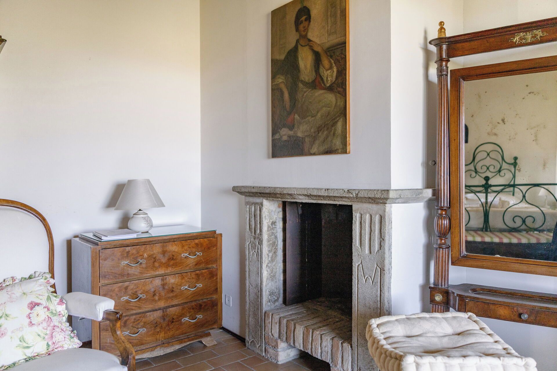 A room with a wooden dresser, a lamp, a painting, a fireplace, and a bed with a headboard.