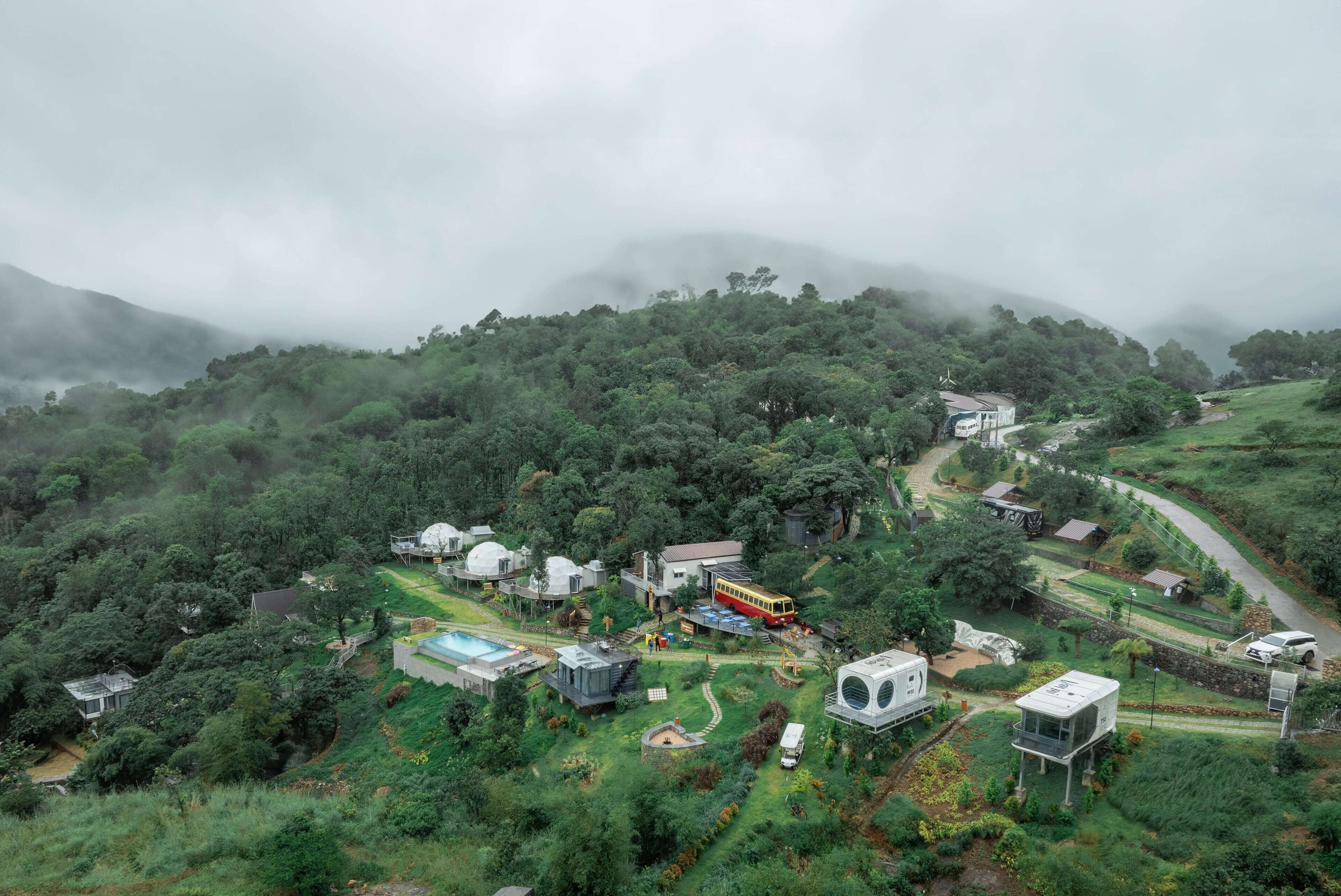 A lush, green landscape with a variety of buildings, including a large dome structure, a bus, and several smaller houses, surrounded by dense forest and misty mountains.