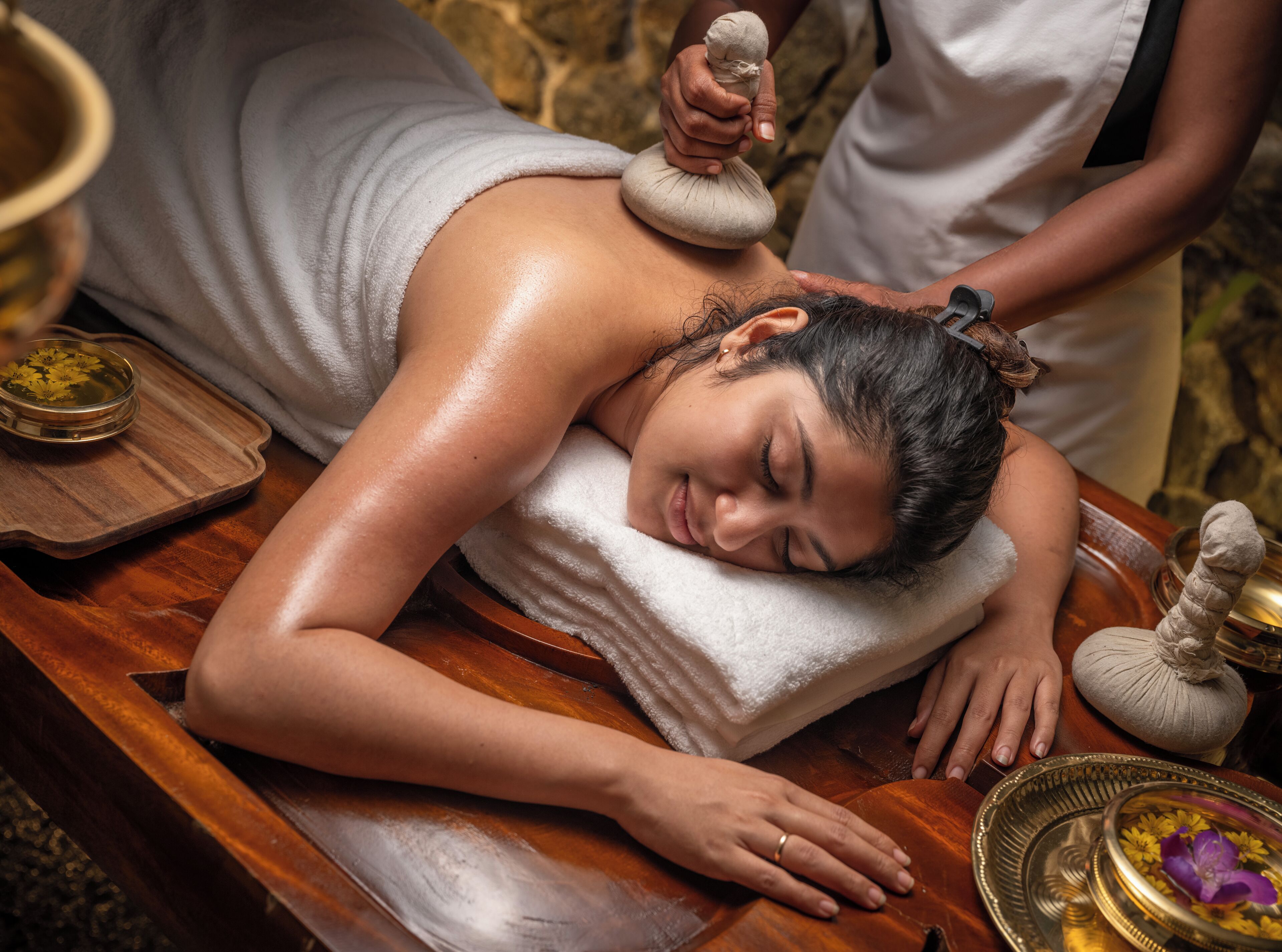A person receiving a massage with a stone on their back, a bowl of flowers, and a wooden tray.