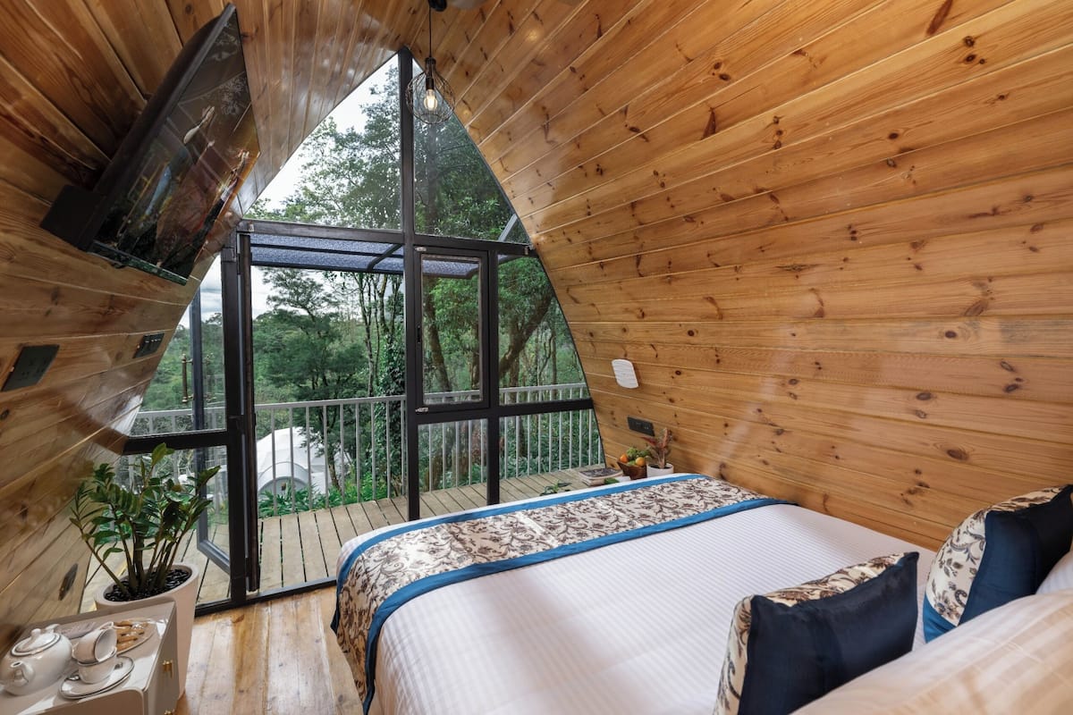 A cozy wooden cabin bedroom with a large window, a bed with patterned bedding, and a small table with cups and a plant.
