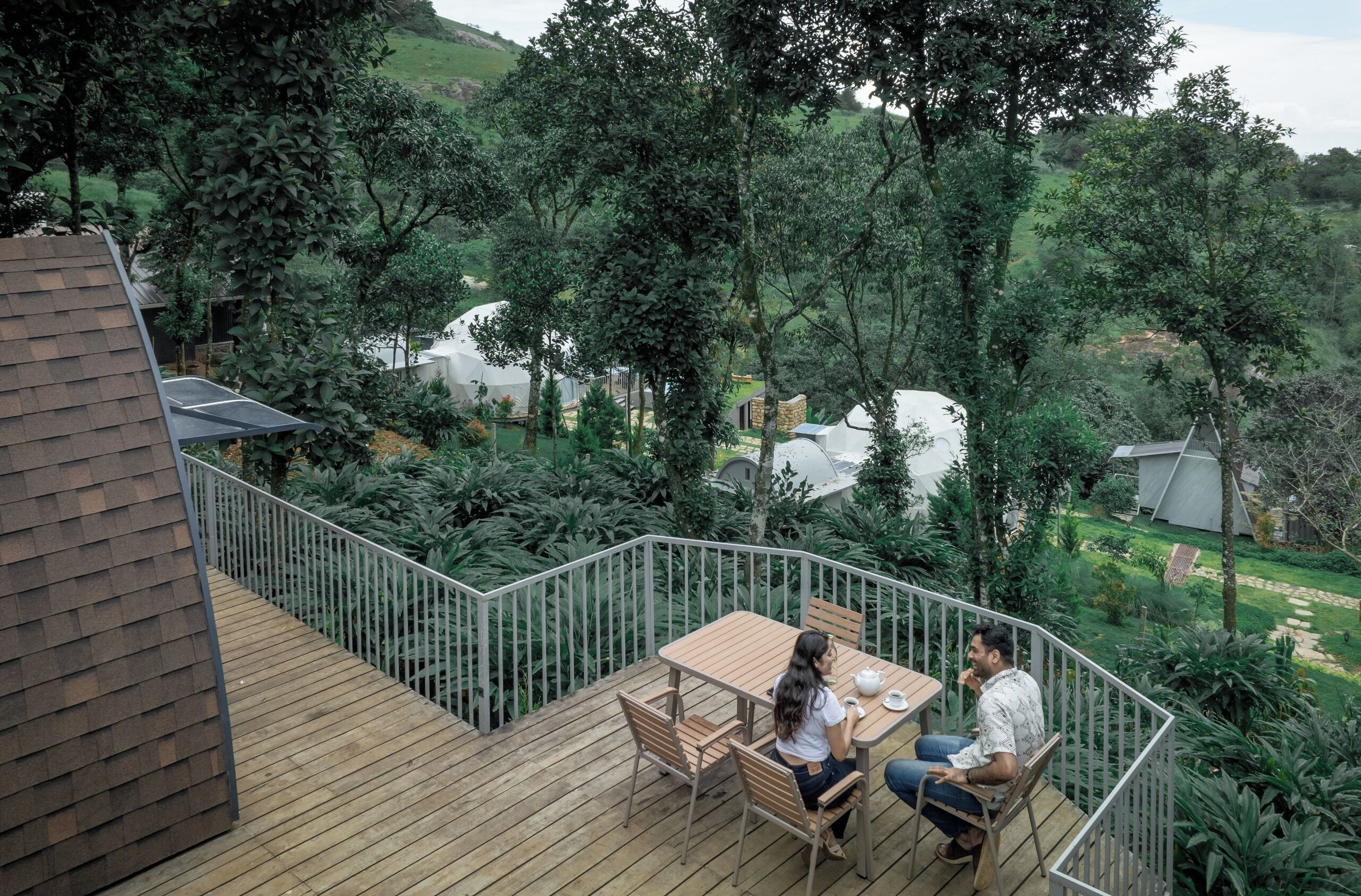 Terraza de madera con mesa y sillas, rodeada por una barandilla metálica, con vistas a un jardín exuberante y un paisaje montañoso.