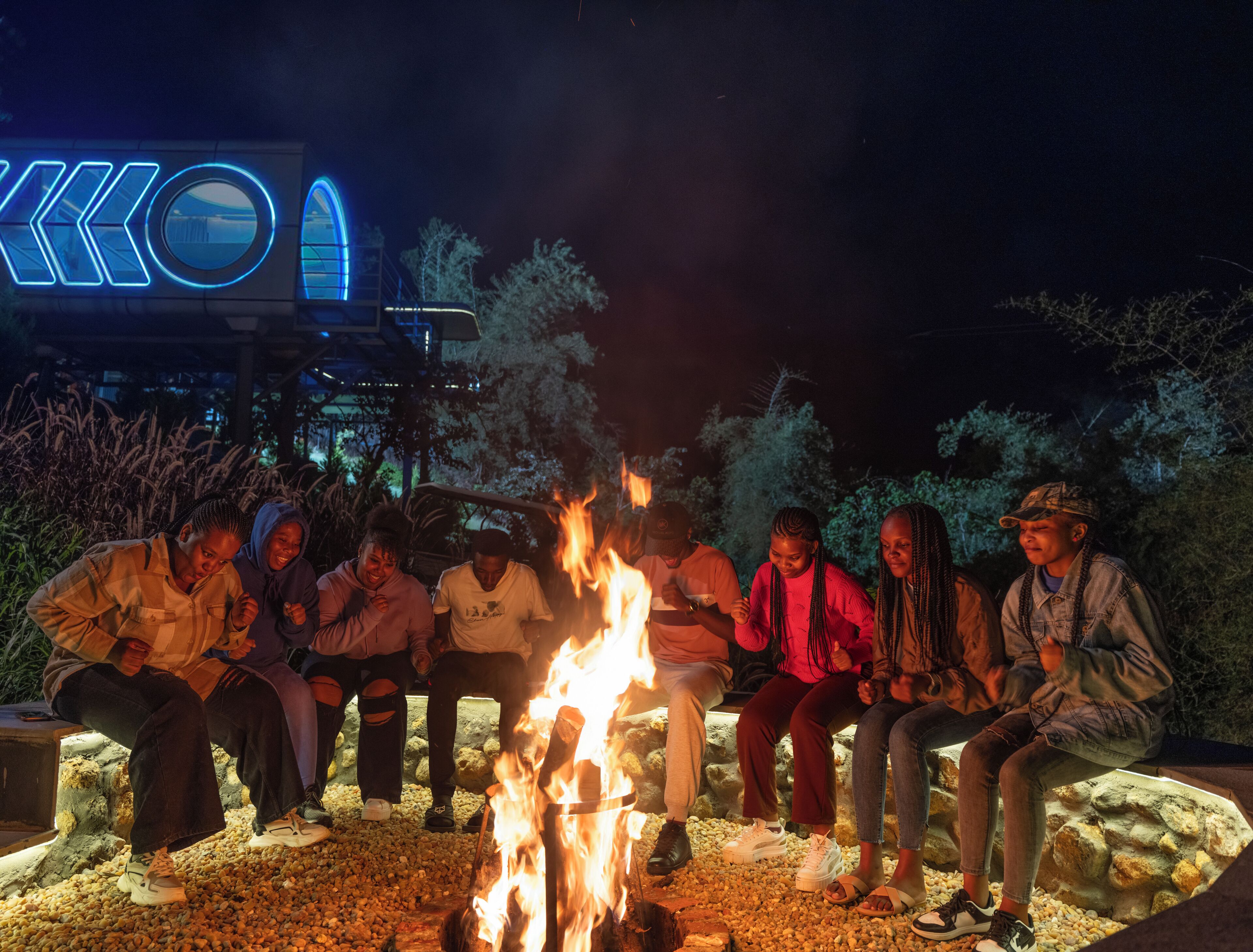 Un groupe de personnes est assis autour d’un feu de camp la nuit, avec un bâtiment en arrière-plan.