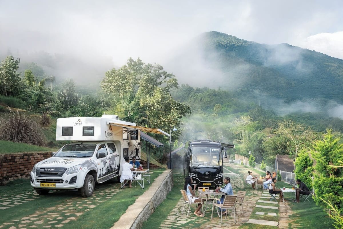 A white RV parked on a grassy area with a black car nearby, surrounded by a garden with tables and chairs, and a mountainous backdrop.