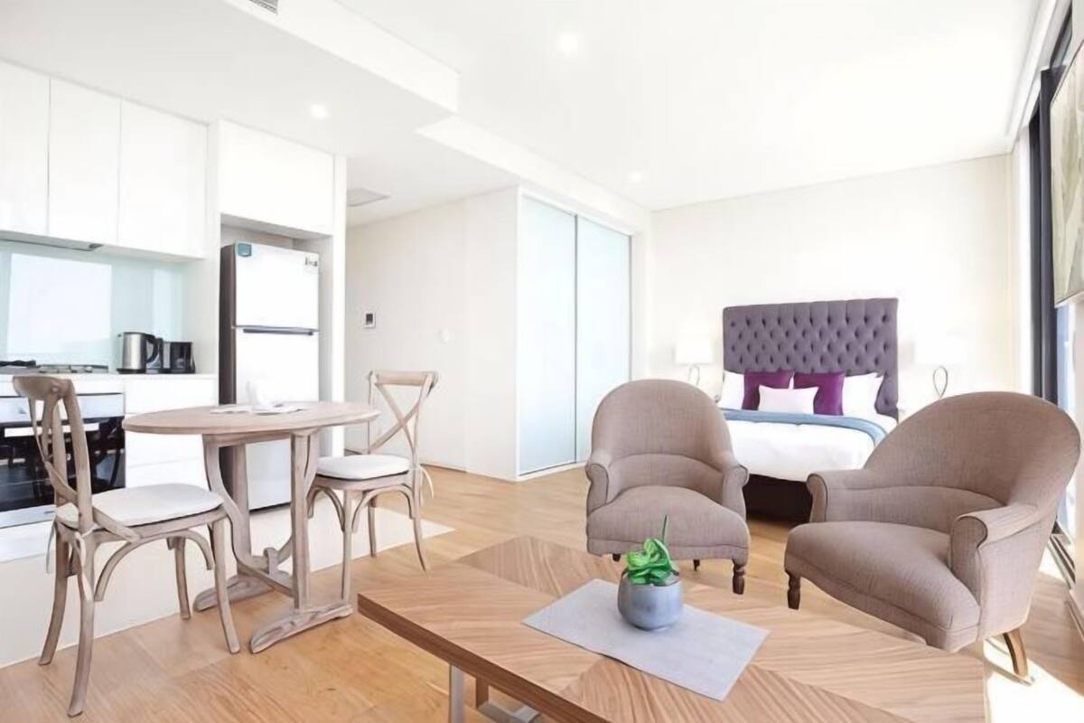 A modern hotel room with a dining area, a bed with a tufted headboard, and two armchairs.