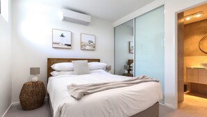 Une chambre moderne avec un grand lit, une table de chevet avec une lampe, un climatiseur fixé au mur et deux tableaux encadrés.