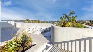 Une terrasse sur le toit, dotée de chaises longues blanches, de jardinières en béton ornées de verdure, et offrant une vue dégagée sur la mer et les collines lointaines.