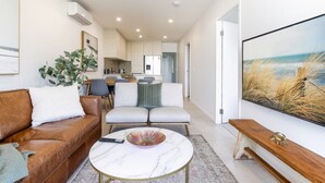 Un salon moderne avec un canapé en cuir marron, un canapé blanc, une table basse en marbre et un grand tableau représentant une scène de plage.