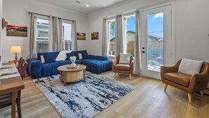 Ein geräumiges Wohnzimmer mit einem blauen Ecksofa, einem braunen Ledersessel, einem runden Couchtisch und einem großen Fenster mit Vorhängen.