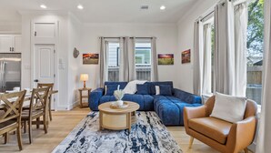 Ein modernes Wohnzimmer mit einem blauen Ecksofa, einem Holztisch und einem braunen Sessel. Es gibt einen Essbereich mit Holzkästen und einem Tisch. Der Raum hat große Fenster mit Vorhängen und Kunstwerken an den Wänden.
